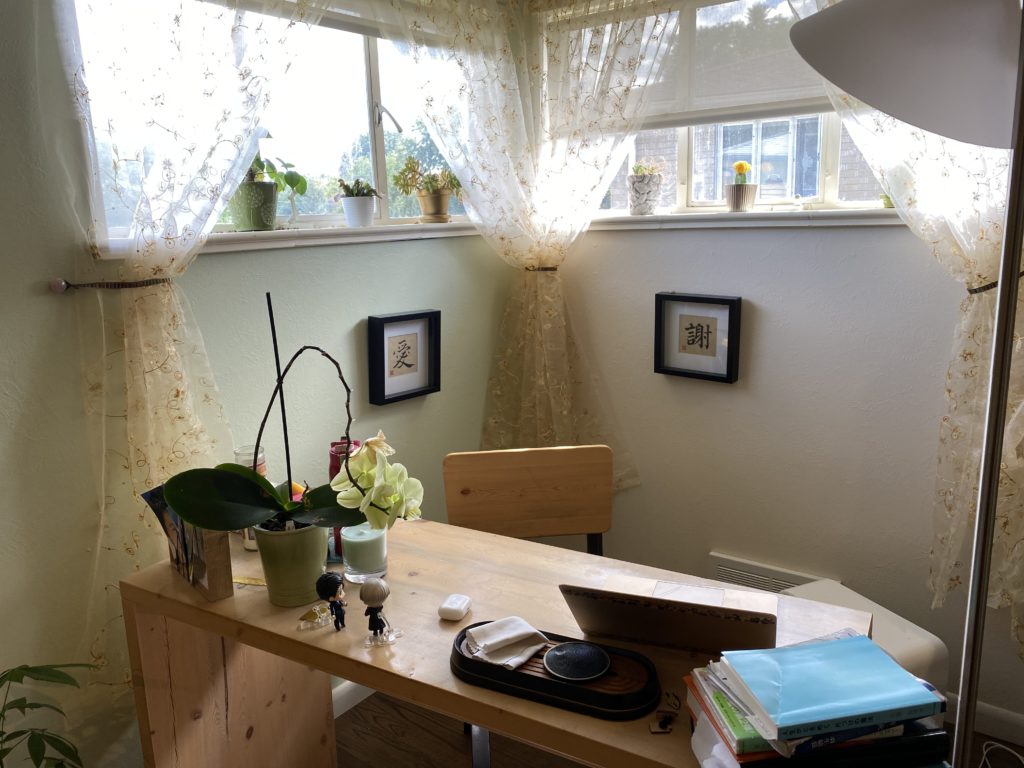 Photograph of my desk: books in the corner, laptop up front, orchid, Yuri on Ice figurines, tea tray on a narrow wooden desk. Behind on the wall are two Chinese characters: 愛 and 謝. Windows are behind, with succulents on the window sill.