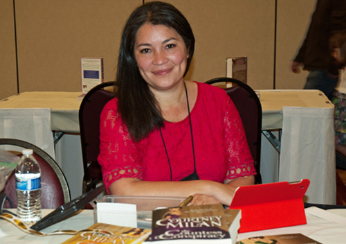 Courtney wearing a fuchsia dress sitting at a table with a book in front of her, ready for signing