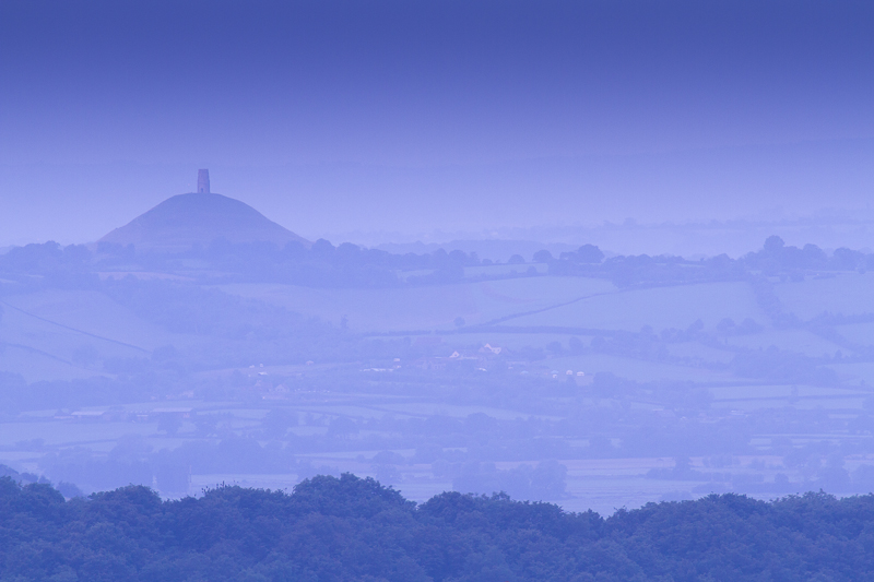 Valley, blue with mist, with Glastonbury Tor, a white monument on a hill, visible