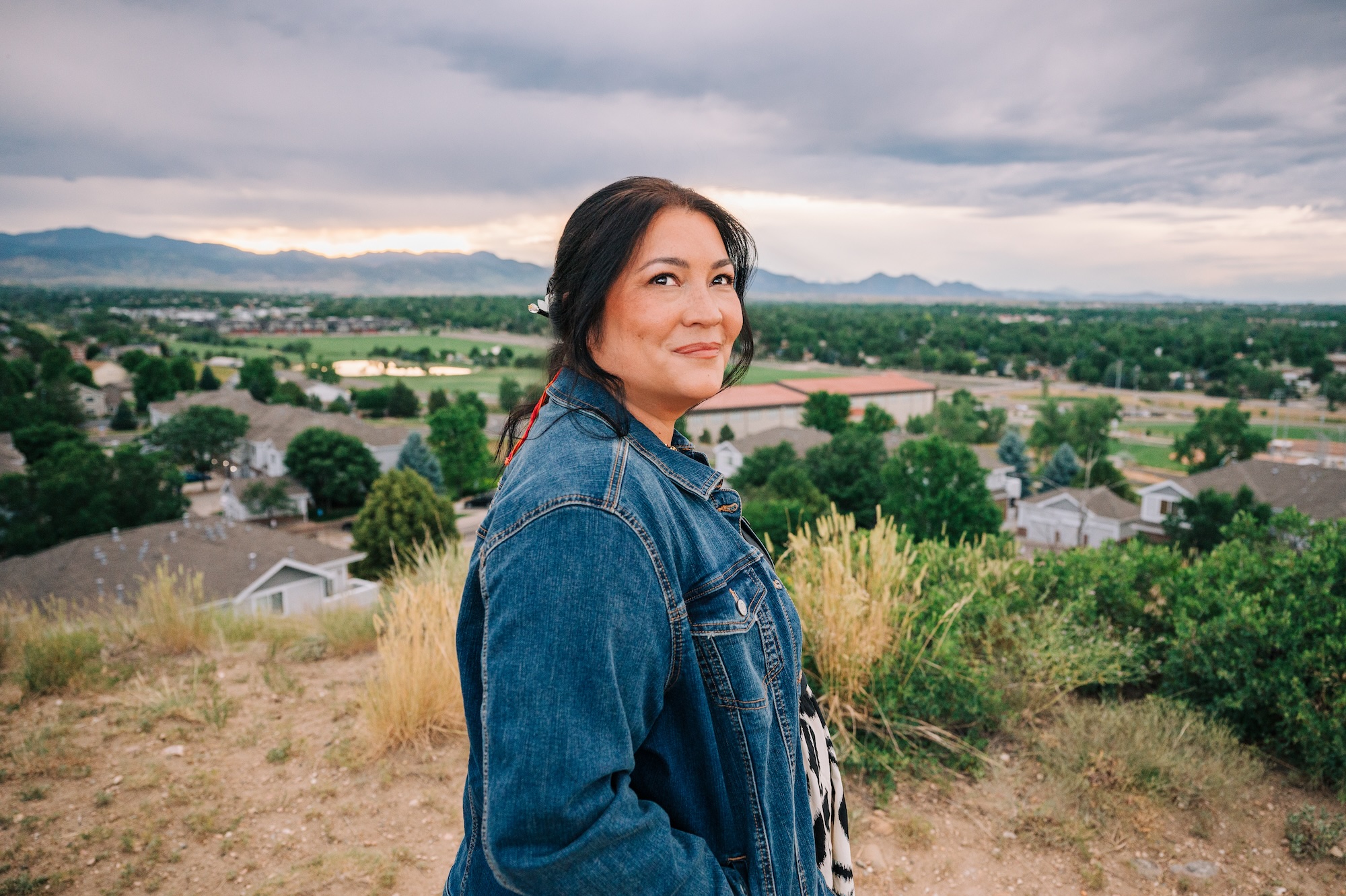 Courtney Milan, standing outside, with the Denver Front range visible behind her, looking up into a stormy sky and smiling